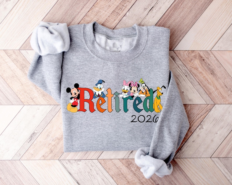 Może przedstawiać: Szara bluza z napisem "Retired" w kolorowych literach, z postaciami Disneya Myszki Miki, Kaczora Donalda, Kaczki Daisy, Myszki Minnie, Goofy'ego i Pluto. Rok "2026" jest wydrukowany poniżej.