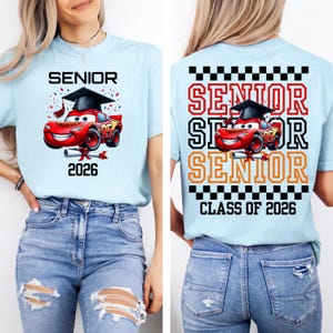 Puede incluir: Camiseta azul claro con tem&aacute;tica de graduaci&oacute;n. La parte delantera presenta la palabra "SENIOR" sobre un coche de dibujos animados rojo con birrete, con el a&ntilde;o "2026" debajo. La parte trasera repite "SENIOR" con el coche.