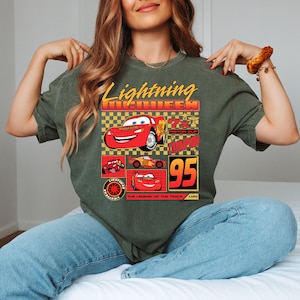 May include: Olive green t-shirt with a graphic of Lightning McQueen from the film Cars. The design includes the text "Lightning McQueen", "95", and other related imagery. The shirt has a classic crew neck.