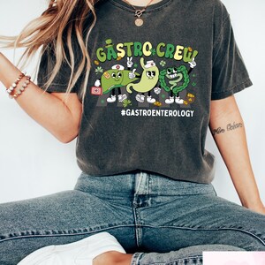 May include: Dark gray t-shirt with a cartoon graphic of three digestive organs labeled "Gastro Crew!" and the hashtag "#Gastroenterology." The design features a liver, stomach, and intestines with smiling faces and medical accessories. The shirt is a casual style.