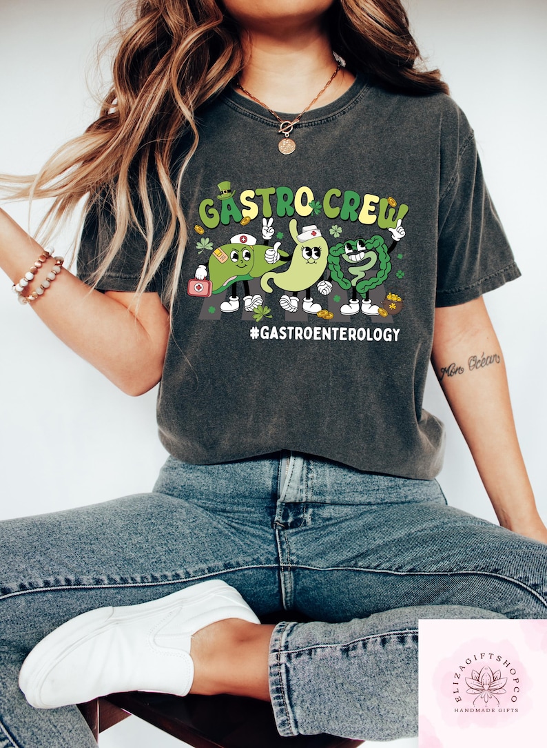 May include: Dark gray t-shirt with a cartoon graphic of three digestive organs labeled "Gastro Crew!" and the hashtag "#Gastroenterology." The design features a liver, stomach, and intestines with smiling faces and medical accessories. The shirt is a casual style.