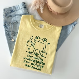 May include: A pale yellow t-shirt featuring a dark green graphic of a frog holding a steaming mug, with the text "currently unavailable for ableist nonsense" printed below. The shirt is displayed alongside light blue denim jeans and a tan hat.