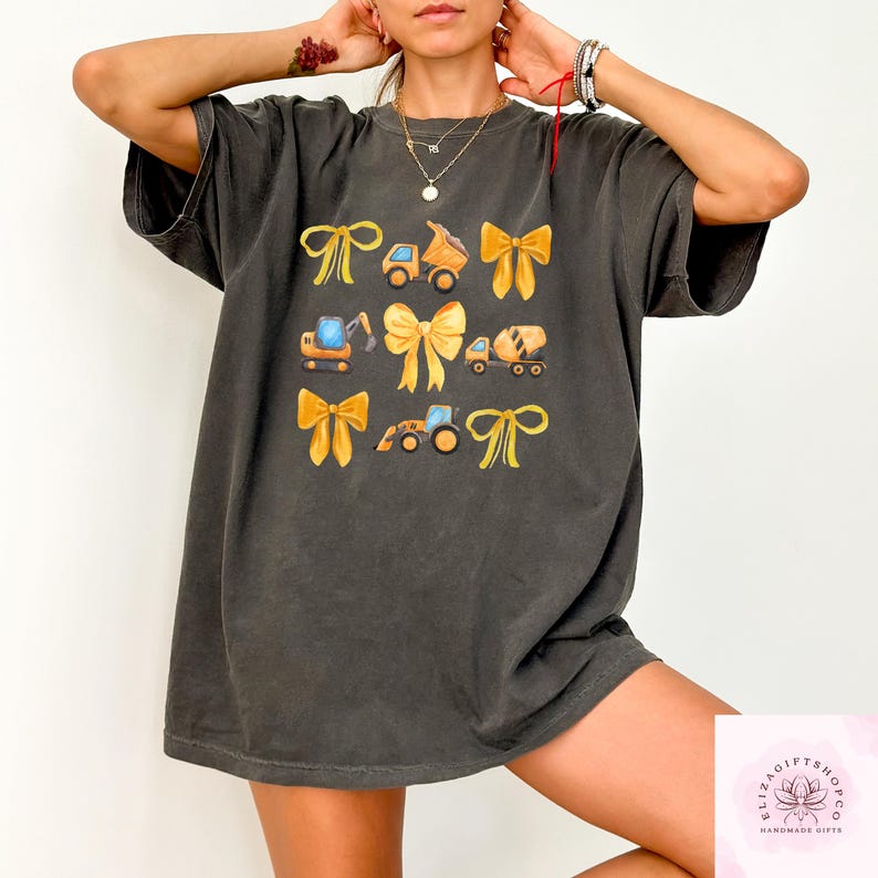 Puede incluir: Camiseta gris oscuro con un dise&ntilde;o de lazos amarillos y veh&iacute;culos de construcci&oacute;n. El dise&ntilde;o incluye un cami&oacute;n volquete, una excavadora y una hormigonera. La camiseta tiene un ajuste c&oacute;modo y holgado.