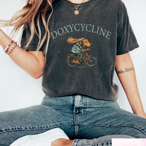 May include: Dark gray t-shirt with the word "DOXYCYCLINE" above an illustration of a dachshund riding a bicycle. The dachshund is wearing a jacket and headphones. The shirt is worn with blue jeans and white sneakers.