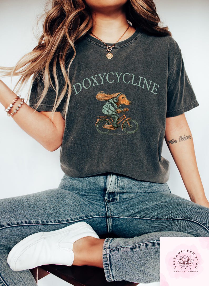 May include: Dark gray t-shirt with the word "DOXYCYCLINE" above an illustration of a dachshund riding a bicycle. The dachshund is wearing a jacket and headphones. The shirt is worn with blue jeans and white sneakers.