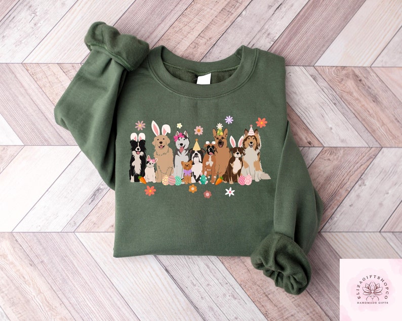 May include: Green sweatshirt with a graphic of a group of dogs wearing Easter hats and bunny ears. The dogs are surrounded by flowers and carrots.
