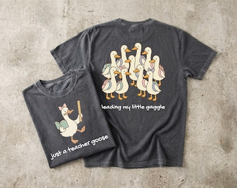 May include: A charcoal gray t-shirt featuring a graphic of a goose holding a book and ruler with the text 'just a teacher goose'. A second t-shirt shows a group of geese with backpacks and the text 'leading my little gaggle' on the back.