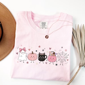 May include: A light pink t-shirt featuring a graphic print of a white ghost with a pink bow, a pink pumpkin, a black cat mug, a pink pumpkin bucket filled with candy, and a spider web, all with smiling faces, surrounded by small pink hearts and stars.