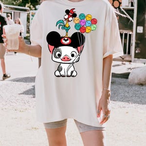 K&ouml;nnte beinhalten: Cremefarbenes &uuml;bergro&szlig;es T-Shirt mit einem Cartoon-Schweinecharakter mit Mickey Mouse-Ohren, einem Hahn und bunten Luftballons. Die Grafik ist in Schwarz, Wei&szlig;, Rot, Rosa, Gelb, Blau und Gr&uuml;n gehalten. Das Shirt ist ein l&auml;ssiger, bequemer Stil.