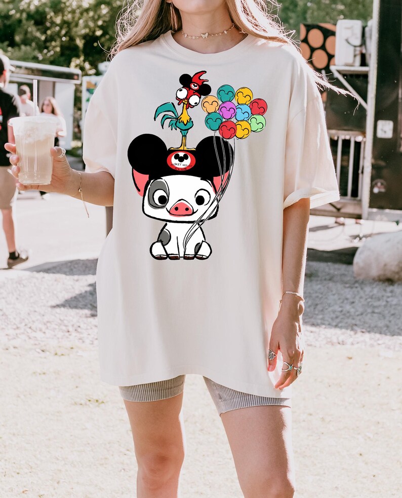 K&ouml;nnte beinhalten: Cremefarbenes &uuml;bergro&szlig;es T-Shirt mit einem Cartoon-Schweinecharakter mit Mickey Mouse-Ohren, einem Hahn und bunten Luftballons. Die Grafik ist in Schwarz, Wei&szlig;, Rot, Rosa, Gelb, Blau und Gr&uuml;n gehalten. Das Shirt ist ein l&auml;ssiger, bequemer Stil.