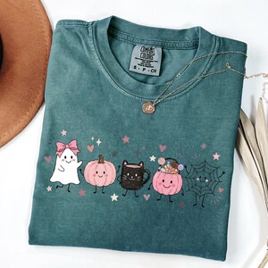 May include: A teal short-sleeved t-shirt featuring a graphic print of a white ghost with a pink bow, a pink pumpkin, a black cat mug, a pink pumpkin bucket filled with candy, and a spider web, all with small pink stars and hearts.