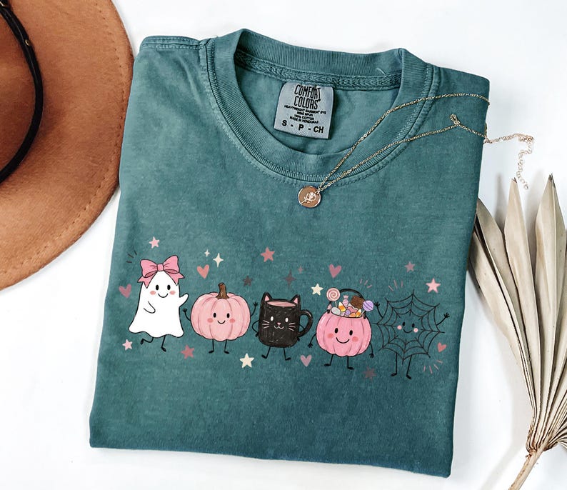 May include: A teal short-sleeved t-shirt featuring a graphic print of a white ghost with a pink bow, a pink pumpkin, a black cat mug, a pink pumpkin bucket filled with candy, and a spider web, all with small pink stars and hearts.