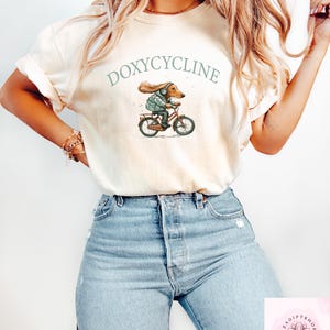 May include: Cream-colored t-shirt featuring the word "DOXYCYCLINE" above an illustration of a dachshund riding a bicycle. The dachshund is wearing a jacket and helmet. The model is wearing light blue jeans.