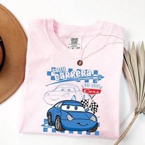 May include: A light pink t-shirt with a blue cartoon car design, the text reads "Sally Carrera" and "Cars Est 2006". The shirt has a crew neck and short sleeves. A gold necklace is draped over the shirt.