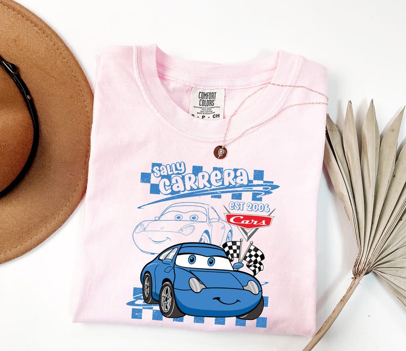 May include: A light pink t-shirt with a blue cartoon car design, the text reads "Sally Carrera" and "Cars Est 2006". The shirt has a crew neck and short sleeves. A gold necklace is draped over the shirt.