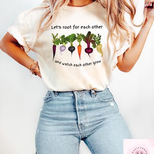 Może przedstawiać: Biała koszulka z napisem "Let's root for each other and watch each other grow" i grafiką kolorowych warzyw.