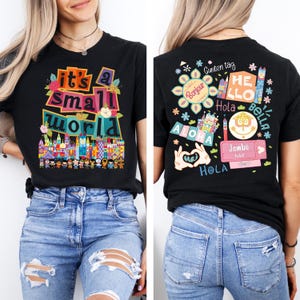K&ouml;nnte beinhalten: Schwarzes T-Shirt mit dem Schriftzug "it's a small world" in bunten Buchstaben und einer skurrilen Illustration einer Stadtlandschaft und verschiedener Figuren. Auf der R&uuml;ckseite befinden sich verschiedene Gr&uuml;&szlig;e und ein Ticket.