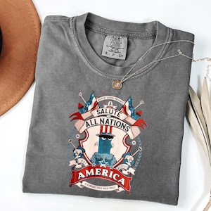 May include: A gray Comfort Colors t-shirt featuring a graphic with the text "A Salute to All Nations, But Mostly America." The design includes a cartoon character, flags, and decorative elements. A silver necklace with a pendant is on the shirt.