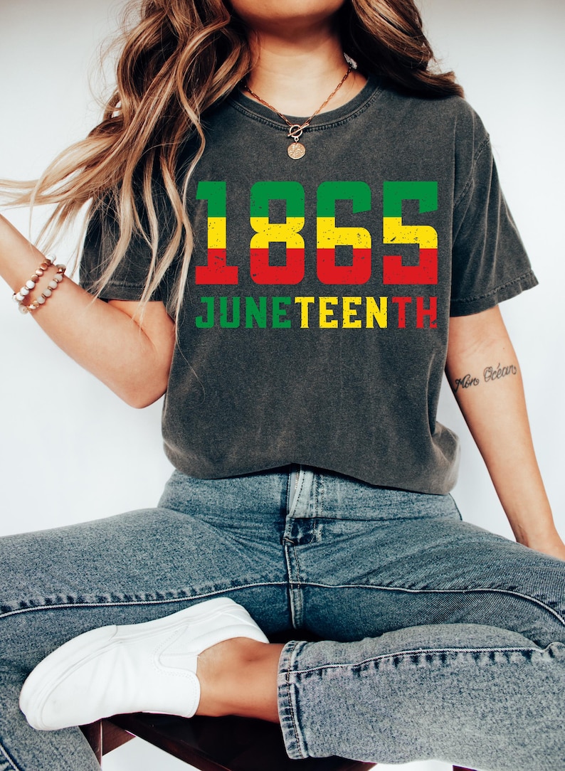 May include: Dark grey t-shirt featuring the text "1865 JUNETEENTH" in green, yellow, and red. The shirt is paired with blue jeans and white trainers. The design commemorates Juneteenth.