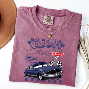 May include: A maroon t-shirt with a graphic of Doc Hudson from the film Cars. The design includes a blue car illustration, the text "Doc Hudson EST 2006 Cars", and checkered flags. The shirt has a crew neck and a Comfort Colors tag.