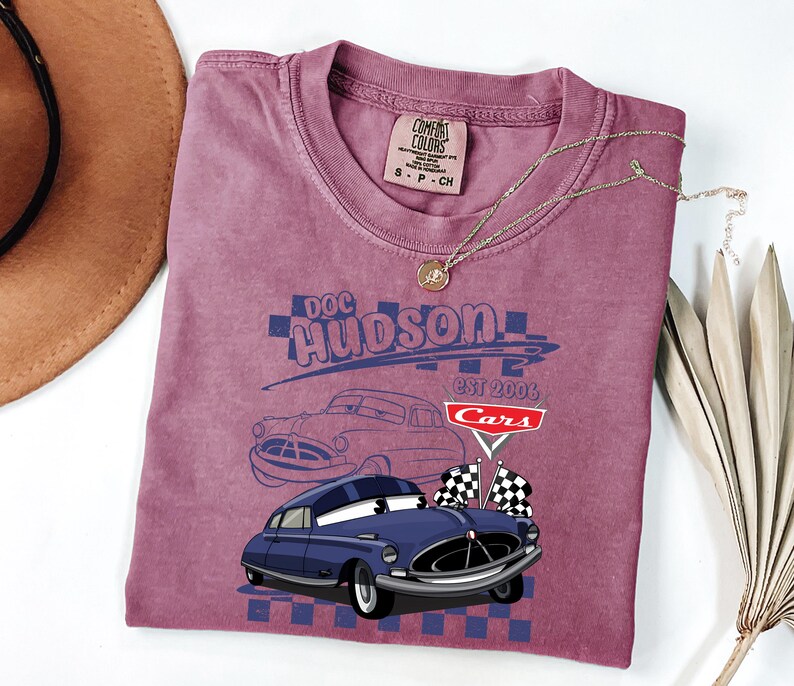 May include: A maroon t-shirt with a graphic of Doc Hudson from the film Cars. The design includes a blue car illustration, the text "Doc Hudson EST 2006 Cars", and checkered flags. The shirt has a crew neck and a Comfort Colors tag.