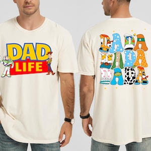 K&ouml;nnte beinhalten: Cremefarbenes T-Shirt mit dem Text "DAD LIFE" in Blau und Gelb, mit Illustrationen von Buzz Lightyear und Woody. Auf der R&uuml;ckseite des Shirts steht "DADA" in bunten, gemusterten Buchstaben. Ein l&auml;ssiges, lustiges Design.