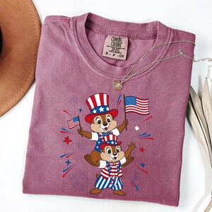 May include: A mauve Comfort Colors t-shirt featuring a patriotic graphic of two chipmunks wearing Uncle Sam hats and holding American flags. The design includes red, white, and blue fireworks and stars. A gold necklace is draped across the shirt.