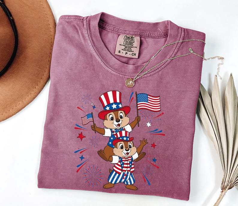 May include: A mauve Comfort Colors t-shirt featuring a patriotic graphic of two chipmunks wearing Uncle Sam hats and holding American flags. The design includes red, white, and blue fireworks and stars. A gold necklace is draped across the shirt.