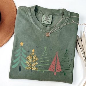 Può includere: T-shirt Comfort Colors verde oliva con una grafica di alberi di Natale stilizzati in verde, giallo e rosso. La maglietta ha la scritta "find things find time" stampata. Una collana d'oro con un ciondolo è sulla maglietta.