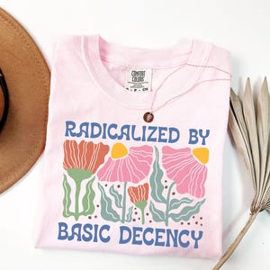 以下が含まれることがあります： 「RADICALIZED BY BASIC DECENCY」の青い文字が入った、薄いピンク色のTシャツ。ピンク、オレンジ、黄色の花柄のデザイン。金のネックレスと茶色の帽子が見えます。