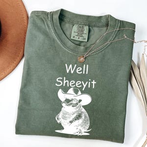 May include: An olive green t-shirt featuring the words "Well Sheeyit" and a graphic of an armadillo in a cowboy hat. A gold necklace and a brown hat are also visible.