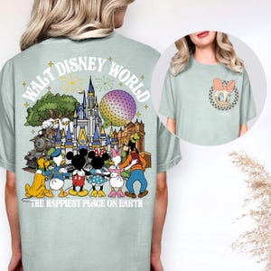 May include: A light green t-shirt featuring a colorful graphic of Disney characters and landmarks, including a train, castle, and Spaceship Earth. The text "WALT DISNEY WORLD" arches across the top, with "THE HAPPIEST PLACE ON EARTH" below. A smaller image shows a Daisy Duck graphic.