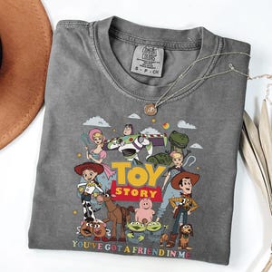 Może przedstawiać: Szara koszulka z kolorową grafiką postaci z filmu "Toy Story", w tym Woody'ego i Buzza Astrala. Projekt zawiera tekst "TOY STORY" i "YOU'VE GOT A FRIEND IN ME". Srebrny naszyjnik jest na koszulce. Rozmiar S-P-CH.