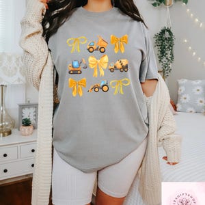 Puede incluir: Camiseta gris con un dise&ntilde;o de lazos amarillos y veh&iacute;culos de construcci&oacute;n. Los veh&iacute;culos incluyen un cami&oacute;n volquete, una excavadora, una hormigonera y un tractor. La camiseta es de estilo casual y c&oacute;modo.