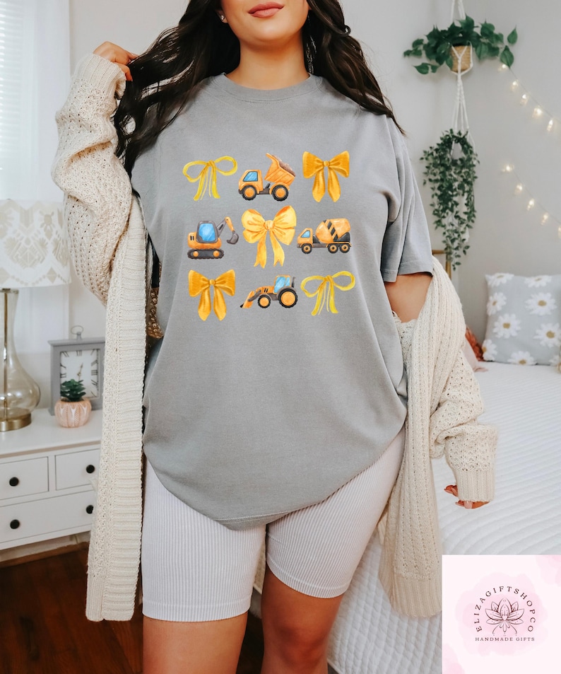 Puede incluir: Camiseta gris con un dise&ntilde;o de lazos amarillos y veh&iacute;culos de construcci&oacute;n. Los veh&iacute;culos incluyen un cami&oacute;n volquete, una excavadora, una hormigonera y un tractor. La camiseta es de estilo casual y c&oacute;modo.