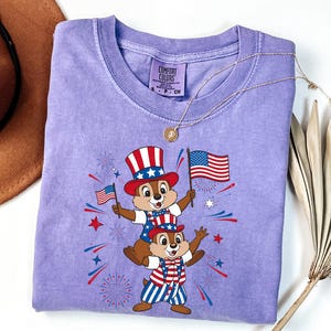 May include: A light purple t-shirt featuring a graphic of two chipmunks dressed in patriotic attire, holding American flags. The chipmunks are wearing Uncle Sam hats and red, white, and blue outfits. The shirt has the text "COMFORT COLORS" on the tag.