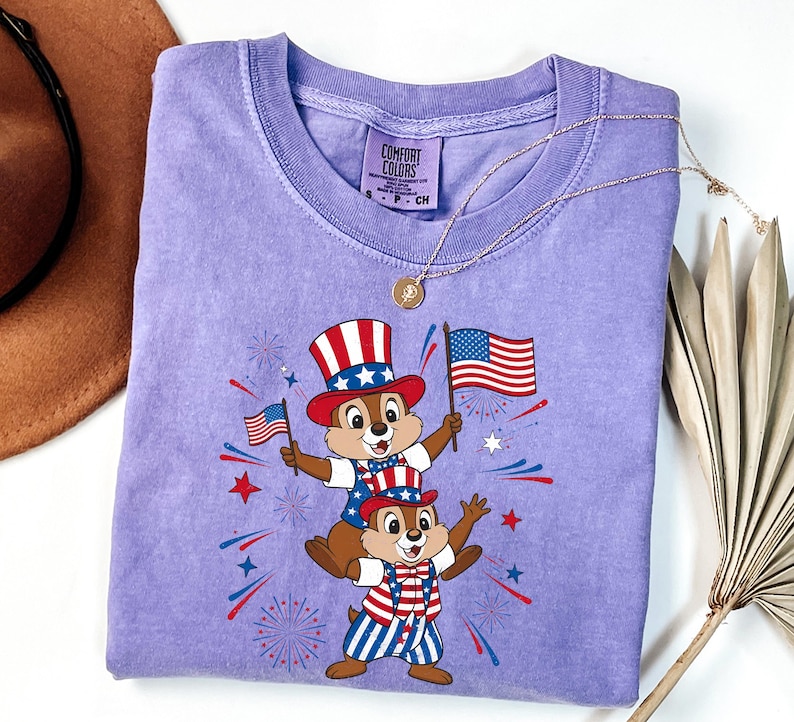 May include: A light purple t-shirt featuring a graphic of two chipmunks dressed in patriotic attire, holding American flags. The chipmunks are wearing Uncle Sam hats and red, white, and blue outfits. The shirt has the text "COMFORT COLORS" on the tag.