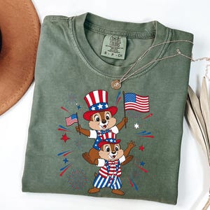 May include: Sage green Comfort Colors t-shirt featuring a patriotic graphic of two chipmunks wearing red, white, and blue outfits and holding American flags. The design includes fireworks and stars, celebrating Independence Day.