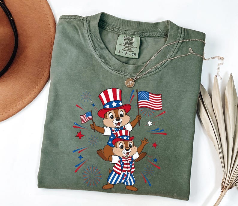 May include: Sage green Comfort Colors t-shirt featuring a patriotic graphic of two chipmunks wearing red, white, and blue outfits and holding American flags. The design includes fireworks and stars, celebrating Independence Day.