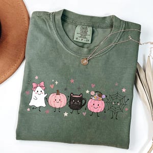 May include: An olive green short-sleeve t-shirt featuring a graphic print of a white ghost with a pink bow, a pink pumpkin, a black cat mug, a pink pumpkin bucket filled with candy, and a spider web, all accented with small pink stars.