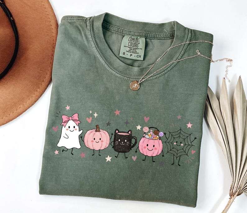 May include: An olive green short-sleeve t-shirt featuring a graphic print of a white ghost with a pink bow, a pink pumpkin, a black cat mug, a pink pumpkin bucket filled with candy, and a spider web, all accented with small pink stars.