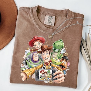 Może przedstawiać: Brązowa koszulka Comfort Colors z grafiką postaci z filmu Toy Story robiących sobie selfie. Projekt zawiera Woody'ego, Buzza Astrala, Jessie, Rexa i inne postacie. Widoczny jest r&oacute;wnież srebrny naszyjnik z wisiorkiem.