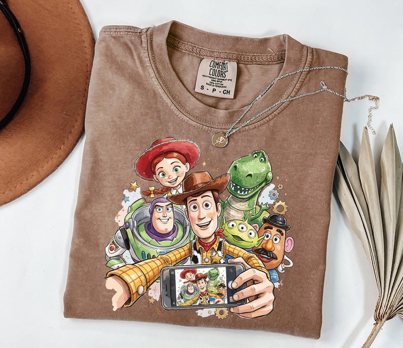 Może przedstawiać: Brązowa koszulka Comfort Colors z grafiką postaci z filmu Toy Story robiących sobie selfie. Projekt zawiera Woody'ego, Buzza Astrala, Jessie, Rexa i inne postacie. Widoczny jest r&oacute;wnież srebrny naszyjnik z wisiorkiem.