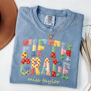 May include: A blue short-sleeve t-shirt with a Comfort Colors label, featuring the text FIFTH GRADE in colorful, patterned fabric letters with apple and gingham prints, and the name miss taylor printed in yellow below the main text.