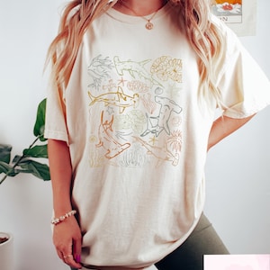 May include: Cream-colored t-shirt featuring a square design with various shark illustrations in shades of brown, green, and gray. The design includes hammerhead sharks, coral, and other marine elements. The shirt is a relaxed fit.
