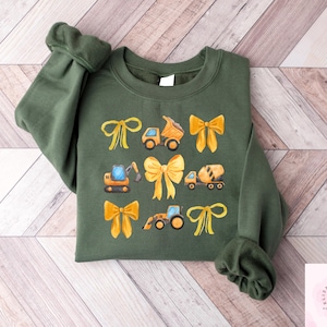 Puede incluir: Una sudadera verde oliva con un dise&ntilde;o de lazos amarillos y veh&iacute;culos de construcci&oacute;n. El dise&ntilde;o incluye un cami&oacute;n volquete, una excavadora, una hormigonera y un tractor. Las mangas est&aacute;n acampanadas.