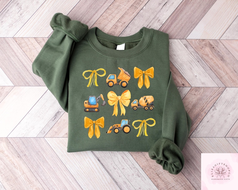 Puede incluir: Una sudadera verde oliva con un dise&ntilde;o de lazos amarillos y veh&iacute;culos de construcci&oacute;n. El dise&ntilde;o incluye un cami&oacute;n volquete, una excavadora, una hormigonera y un tractor. Las mangas est&aacute;n acampanadas.