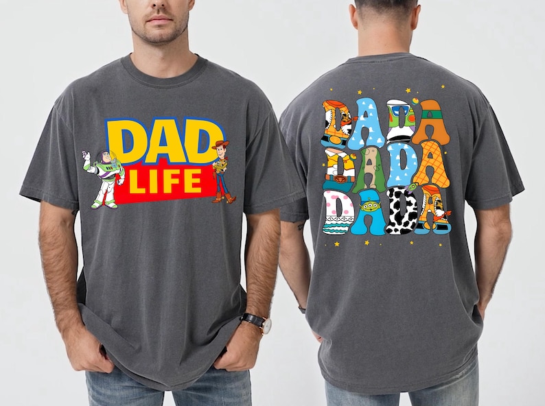 K&ouml;nnte beinhalten: Dunkelgraues T-Shirt mit "DAD LIFE" in Gelb und Rot, mit Buzz Lightyear und Woody. Auf der R&uuml;ckseite des Shirts steht "DADA DADA" in bunten, cartoonartigen Buchstaben. Ein lustiges, thematisches Kleidungsst&uuml;ck.