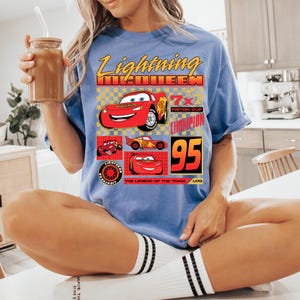 May include: A light blue t-shirt featuring Lightning McQueen from the Cars film. The graphic includes the text "Lightning McQueen", "7x Piston Cup Champion", and the number "95". The shirt has a crew neck and short sleeves.