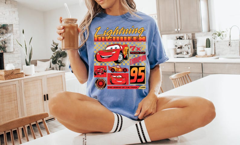 May include: A light blue t-shirt featuring Lightning McQueen from the Cars film. The graphic includes the text "Lightning McQueen", "7x Piston Cup Champion", and the number "95". The shirt has a crew neck and short sleeves.